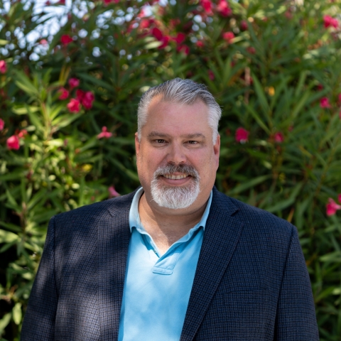 Brad in a light blue collared shirt under a navy blue blazer in front of green bushes with pink flowers. 